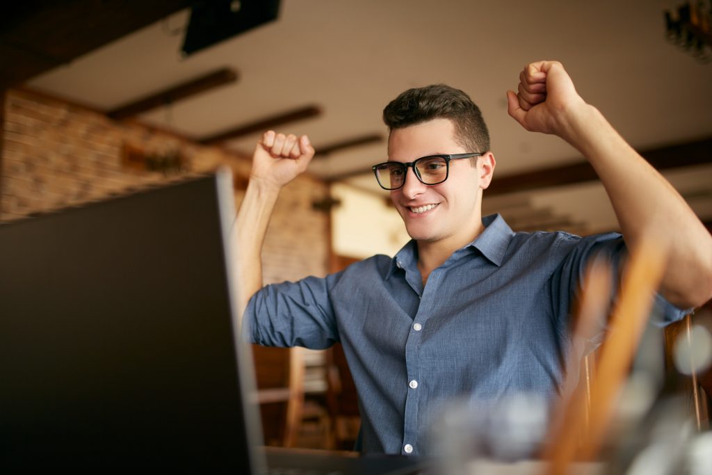 Handsome businessman with laptop having his arms with fists raised, celebrating success. Happy freelancer hipster in glasses finished work on project. Man won a lot of money in lottery prize.