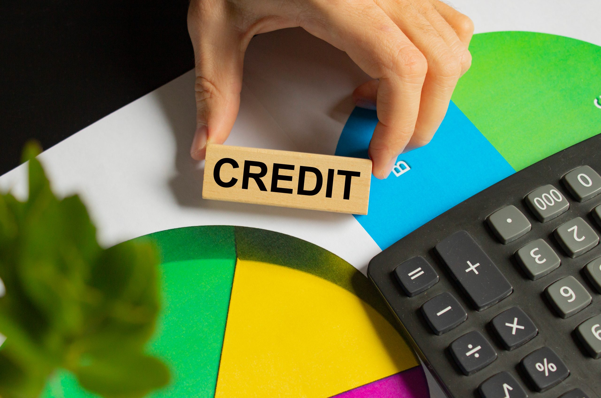 The word credit is written on a wooden block held by a male hand on an office desk with a calculator and color charts.