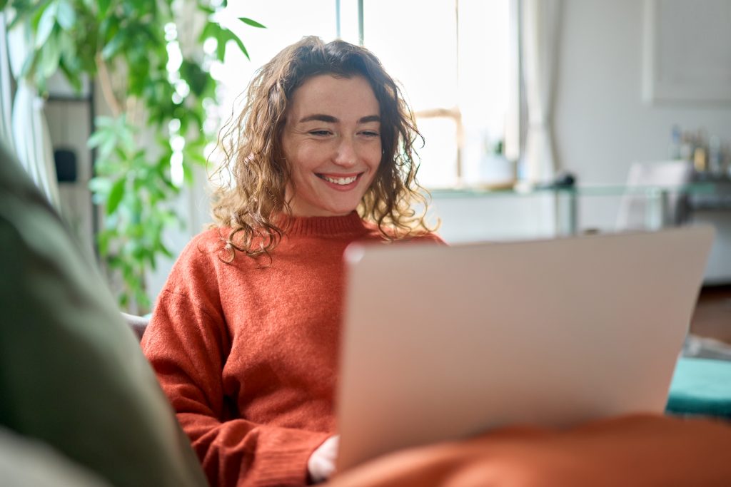 Relaxed happy young woman sitting on sofa using laptop at home surfing.