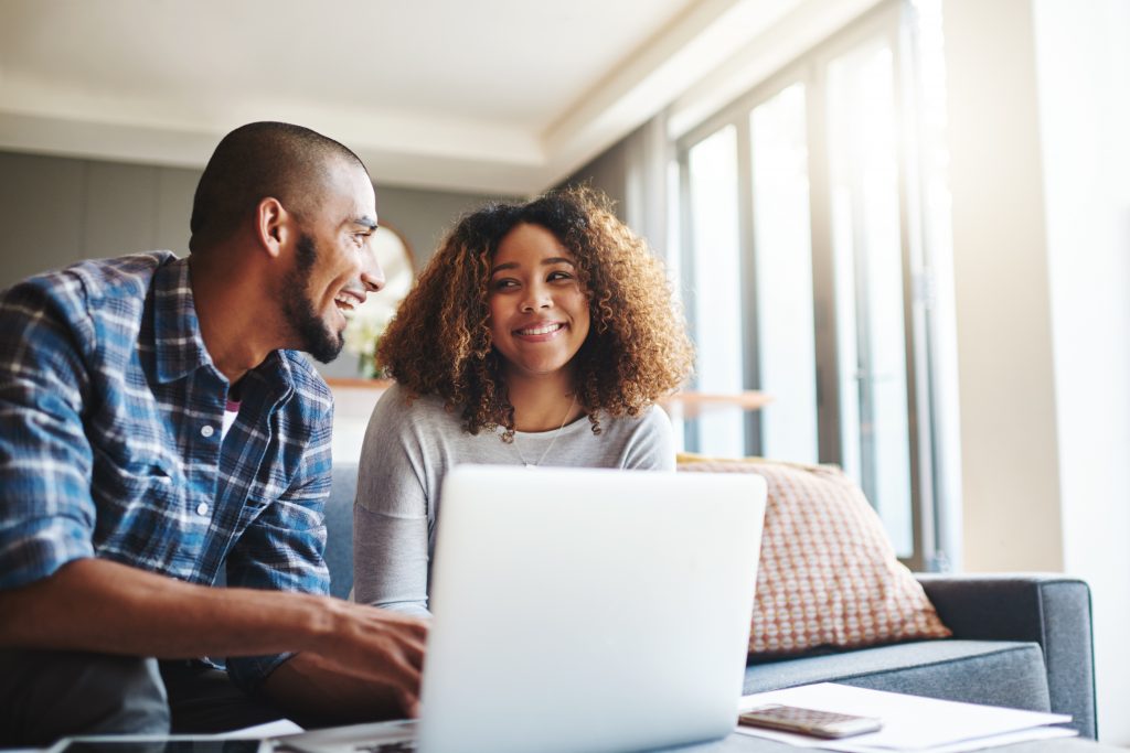 Happy, excited and cheerful couple browsing on laptop and using the internet for payments and calculating home budget while sitting on sofa. Young husband and wife enjoying convenient online banking.