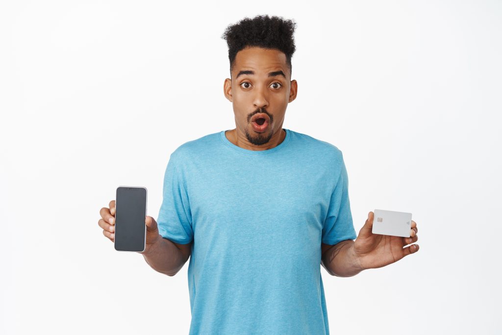 Portrait of excited african american guy showing mobile phone empty screen, credit card, gasping amazed, checking out smartphone shop, internet store discount, white background