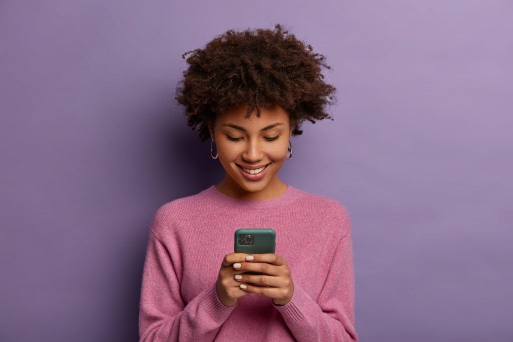 Pretty curly woman holds modern mobile phone, types messages on smartphone device, enjoys online communication, downloads special app for chatting, smiles tenderly, isolated on purple studio wall