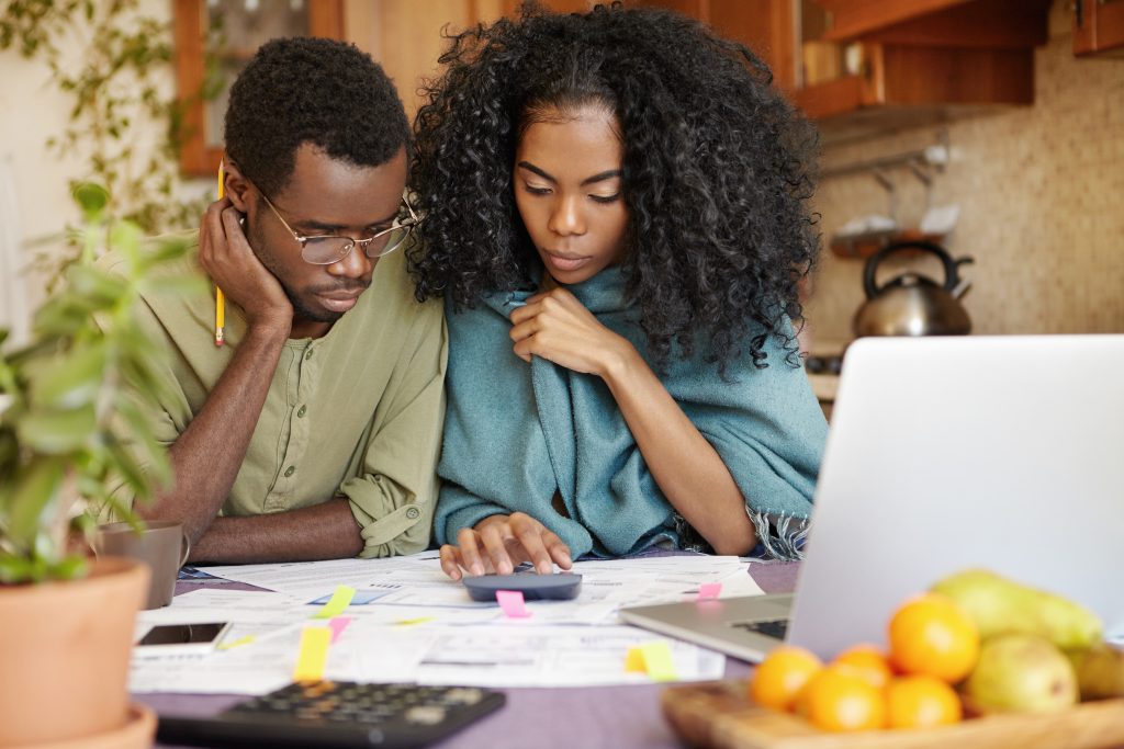 Unhappy and depressed young African-American couple calculating family budget, planning expenses, trying to make both ends meet, not able to pay off all their debts. Financial problem and economy