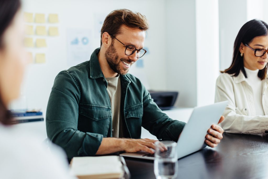 Designer using a laptop during a meeting in an office. Happy business man reading an email while sitting in a boardroom with his colleagues.