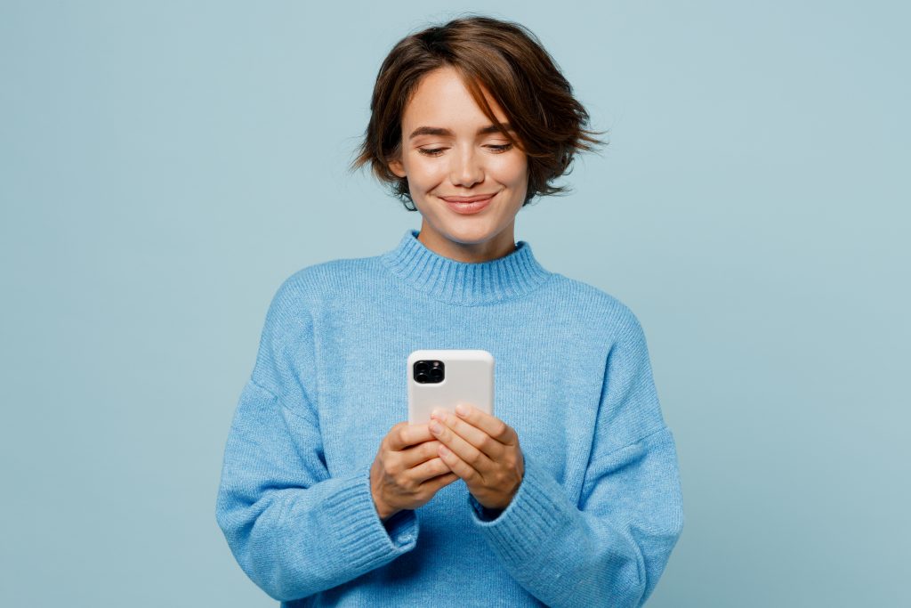 Young smiling happy fun caucasian woman wear knitted sweater hold in hand use mobile cell phone chatting isolated on plain pastel light blue cyan background studio portrait. People lifestyle concept.