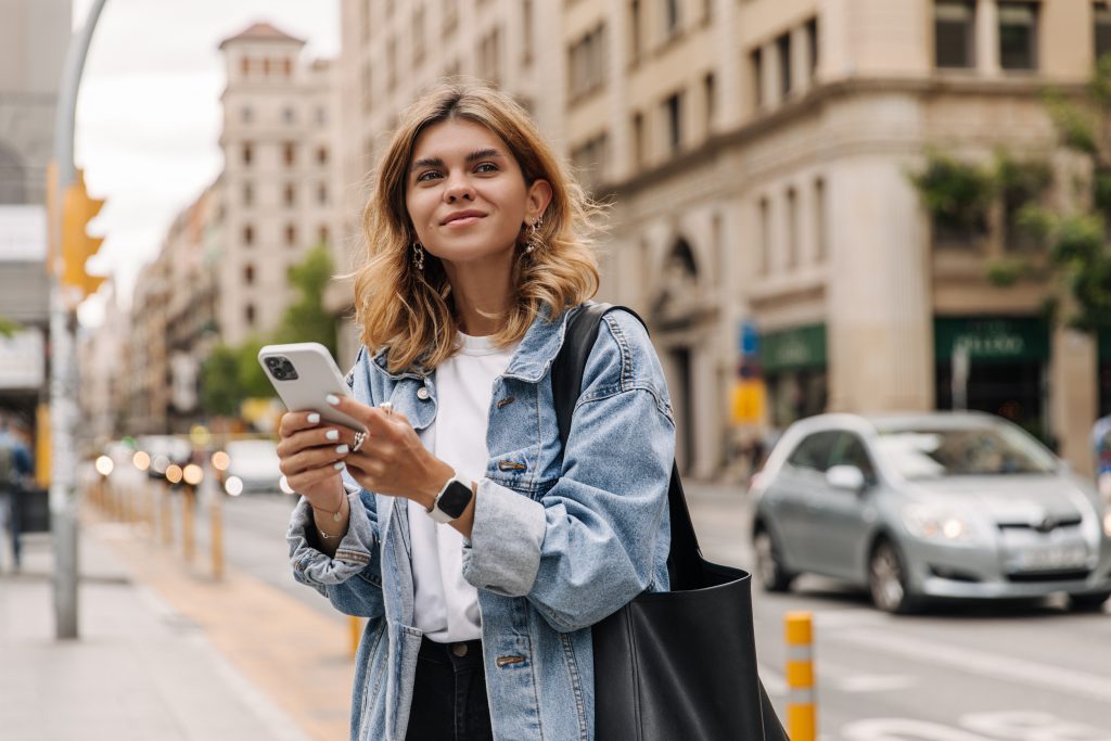 Picture of pretty young woman staying on the street holding phone in hands.