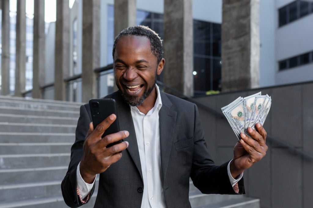 African american man in business suit rejoices, got online small business loan, owner with money cash dollars happy with achievement result, boss holding cash and phone