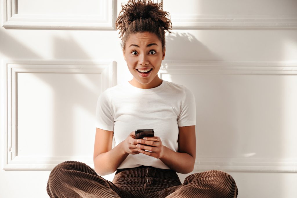 Amazed african american woman holding smartphone. Surprised black girl sitting on floor under sun rays