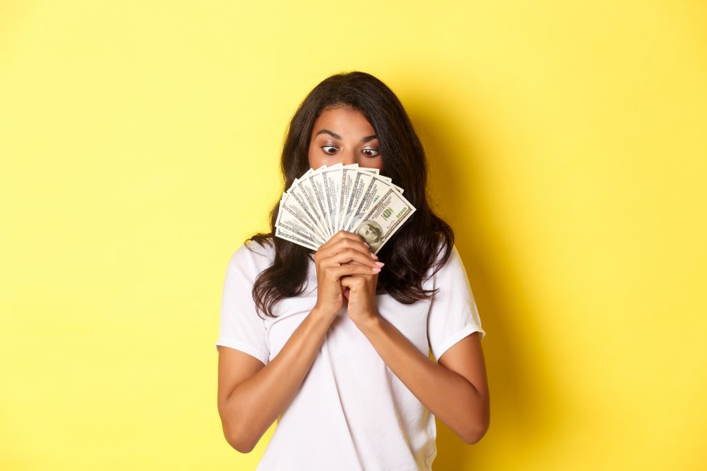 Portrait of lucky african-american girl winning money prize, holding cash and looking amazed, standing over yellow background