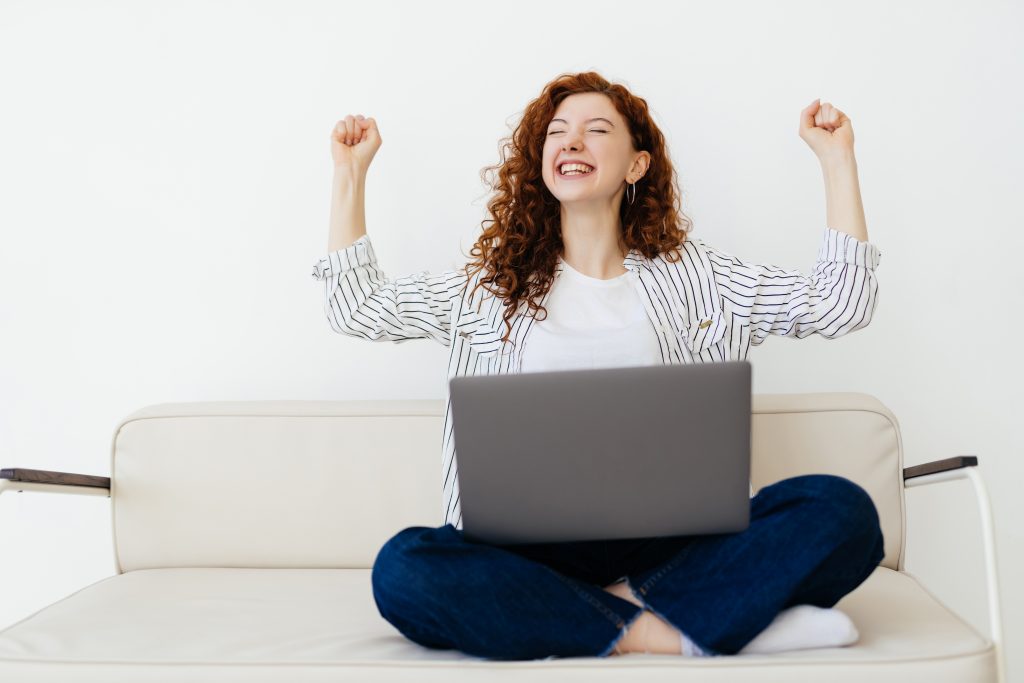 Portrait of a pretty young woman celebrating a success on laptop sit on couch