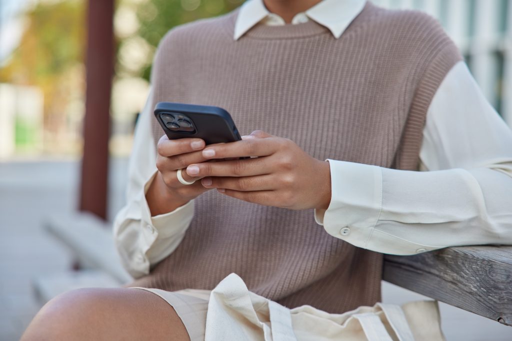 Cropped image of unrecognizable woman wears neat clothes uses mobile phone sends text messages and mails chats onlie sits on wooden bench against blurred background spends free time in street