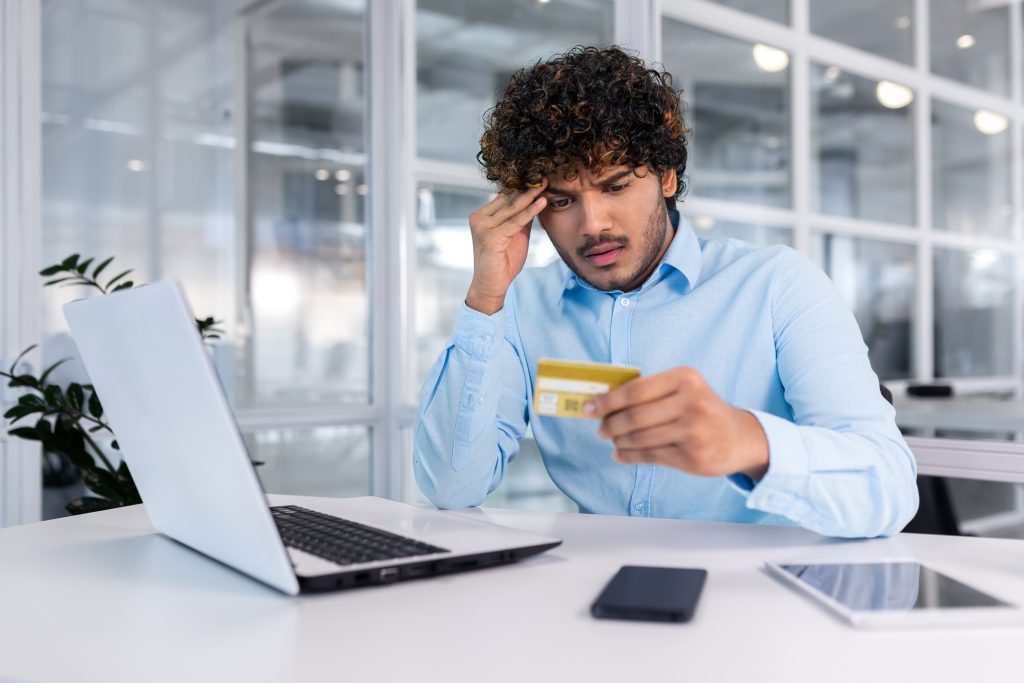 Problems with the account, bankruptcy, etc. A young hispanic man is sitting at the desk in the office, looking worriedly at the credit card, holding his head