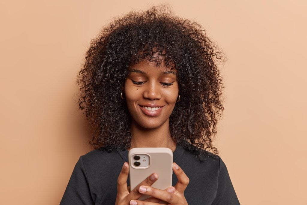 Cheerful woman with Afro hair engages in text messaging on her mobile phone searches for holiday gifts on internet browsing webpages through smartphone app enjoys convenience of virtual shopping