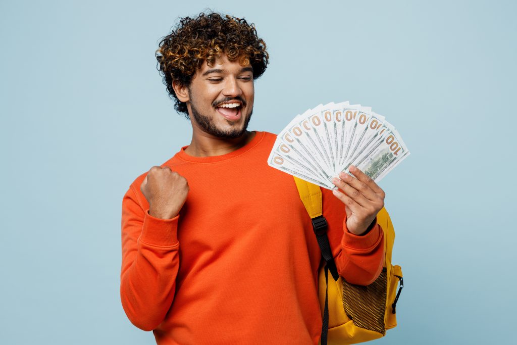 Young teen Indian boy student wears casual clothes backpack bag hold fan of cash money in dollar banknotes, do winner gesture isolated on plain blue background. High school university college concept.