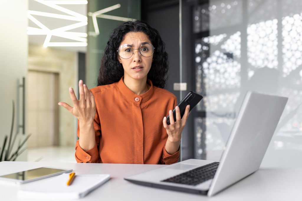 Frustrated and sad business woman with phone in hands looking at camera, latin american woman working with laptop inside modern office building, unclear emotional state of female employee.