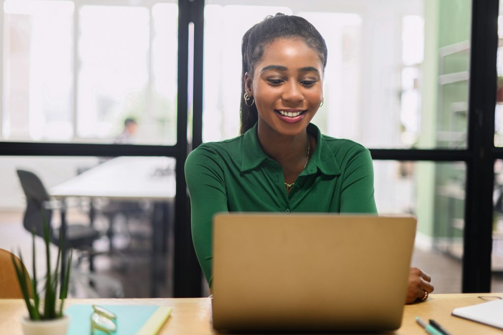 Friendly and positive african-american female employee or student using laptop, multiracial business woman looking at the screen, typing, responding to emails sitting in office