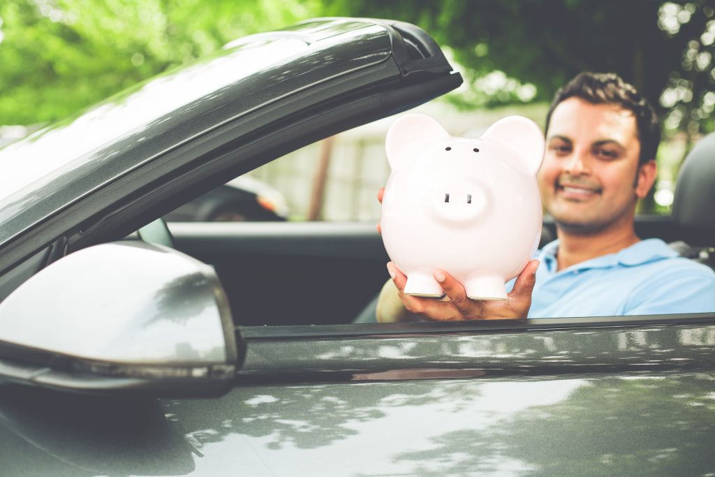 Closeup portrait, handsome young man in blue polo shirt showing pink piggy bank inside new sports car, isolated outdoors background. Happy to have good low apr interest rates, finance rebate deals