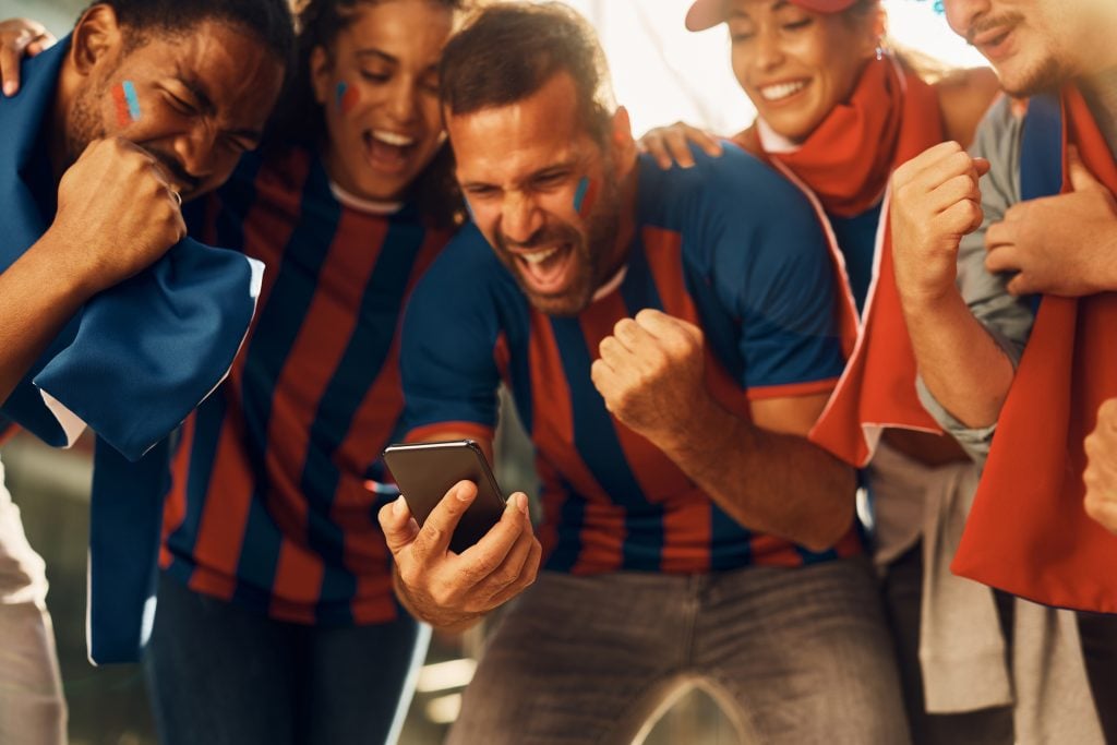 Excited soccer fans celebrating winning goal of their favorite team while watching match on smart phone.
