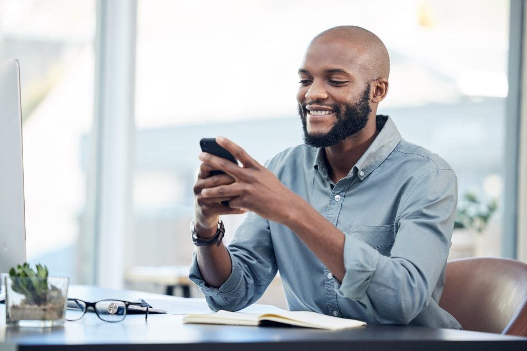 Black man in office, check social media on smartphone and smile at meme, lunch break and communication. Male employee at workplace, using phone and technology, mobile app and contact with chat