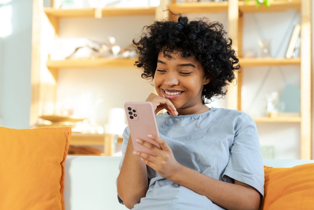 Excited happy young black african american woman holding cell phone laughing feeling joy getting mobile message. Overjoyed girl laughing aloud sitting on couch watching funny video reading news.